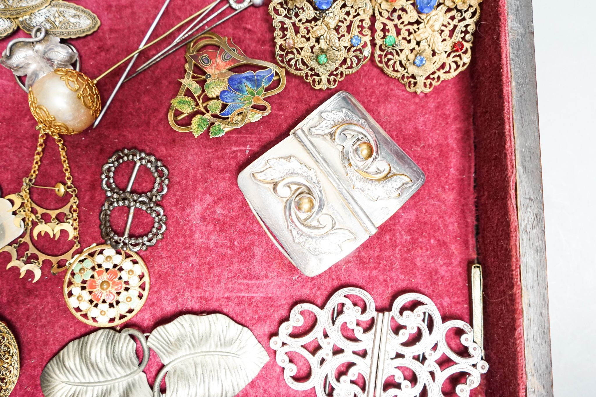 A group of belt buckles, clips and hatpins in a mahogany display case, 30.5cm